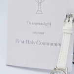 A white gift box with a chalice logo and the words 'First Holy Communion' inscribed on it, accompanied by a white PU strap watch with a cz detail on the strap.