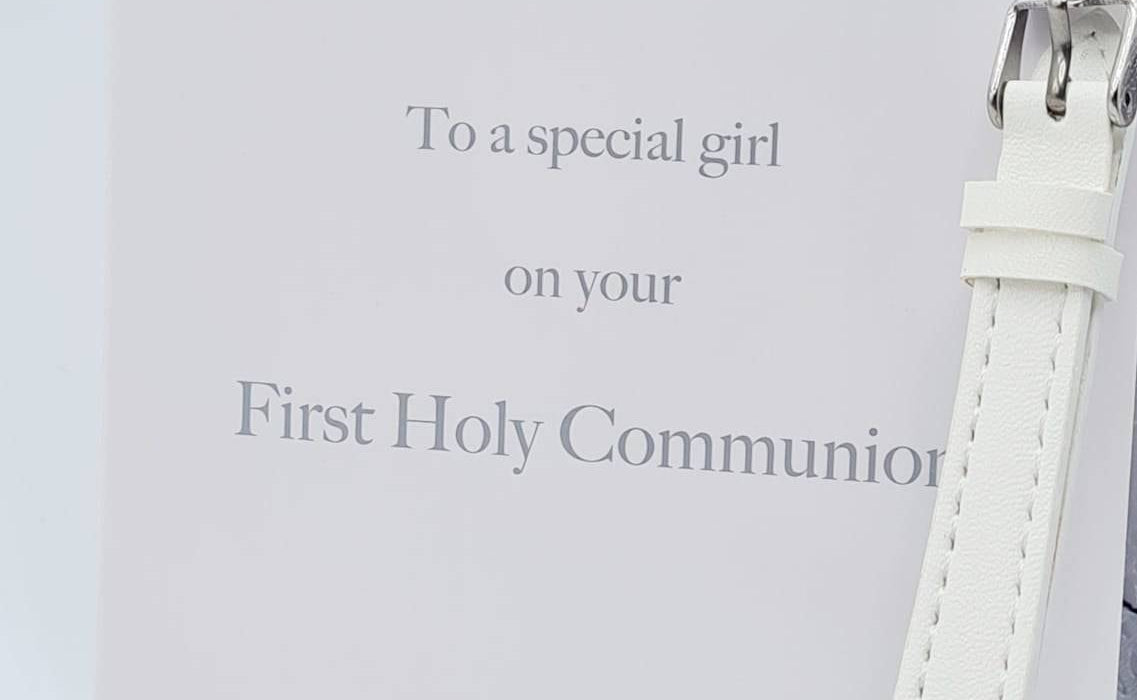 A white gift box with a chalice logo and the words 'First Holy Communion' inscribed on it, accompanied by a white PU strap watch with a cz detail on the strap.
