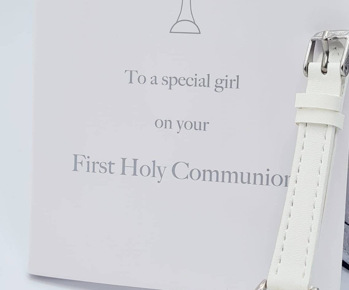 A white gift box with a chalice logo and the words 'First Holy Communion' inscribed on it, accompanied by a white PU strap watch with a cz detail on the strap.